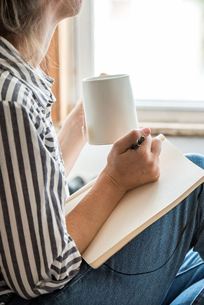 Female looking out window while holding mug in left hand and a pen in right with a journal in her lap. We see her from the chin down. She's wearing a black and white striped button down shirt.