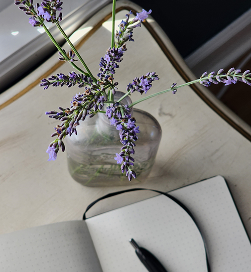 overhead shot of white desk with a open blank journal, pen on the page, and a vase of lavender