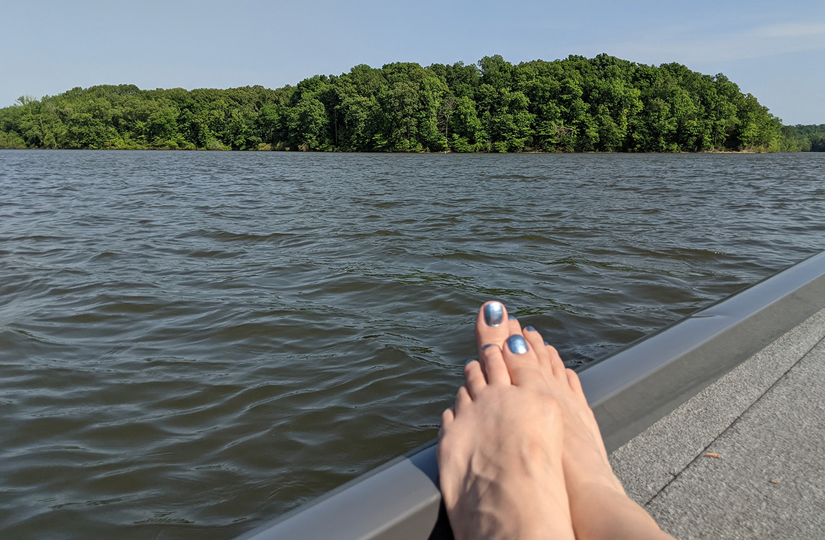 Bare feet resting on a boat's edge that's on a lake with forest shoreline in far distance 