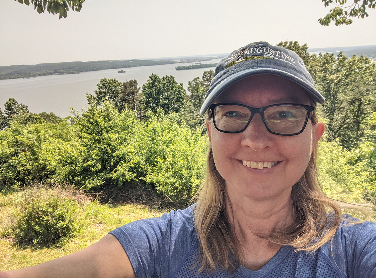 A selfie of the author, Jackie Brown, as she stands on an overlook with the Tennessee River way off in the background, bushes in the middle ground.