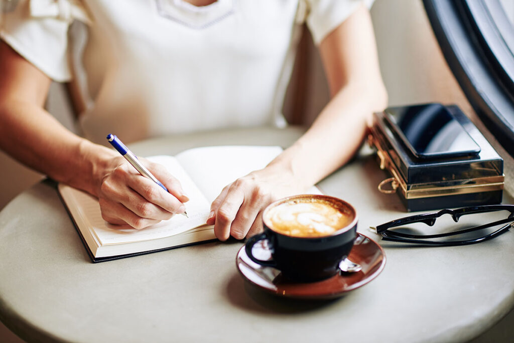 gray table with a black coffee mug, a stack of books and a woman's arms and hands over an open journal